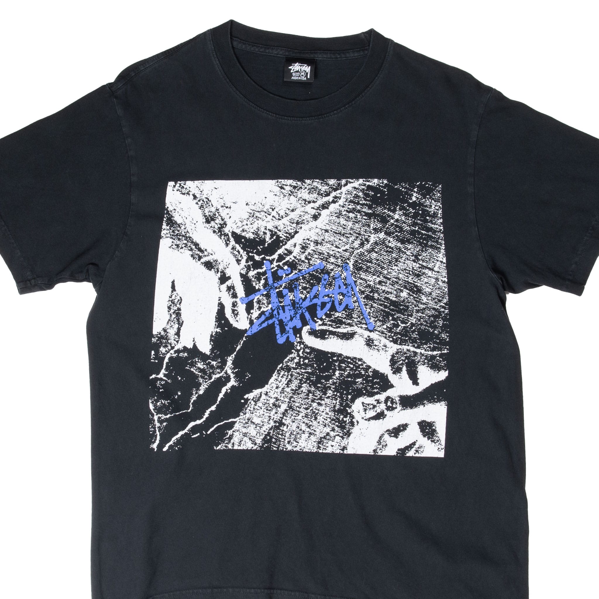 Front upper view of black Stüssy T-shirt with photographic chest graphic and blue script logo