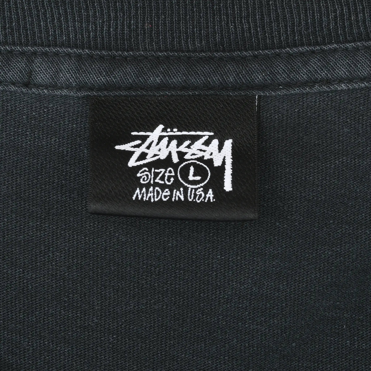 Close-up of black Stüssy neck tag marked size L and Made in U.S.A.