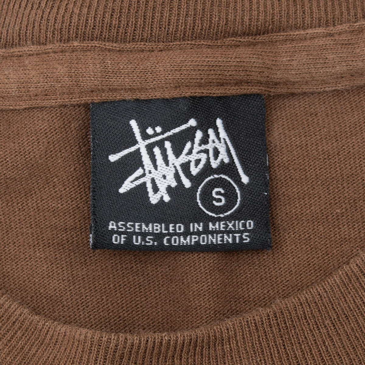 Close-up of black Stussy neck label marked size S inside brown T-shirt