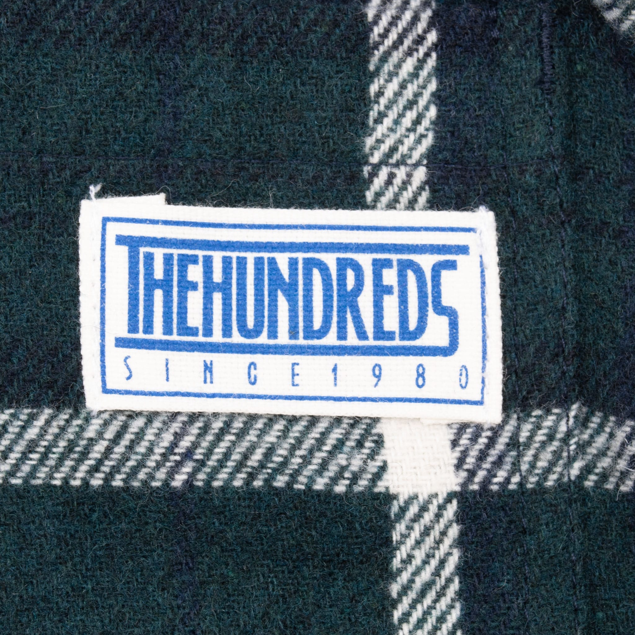 Close-up of The Hundreds Since 1980 chest patch on green plaid fabric