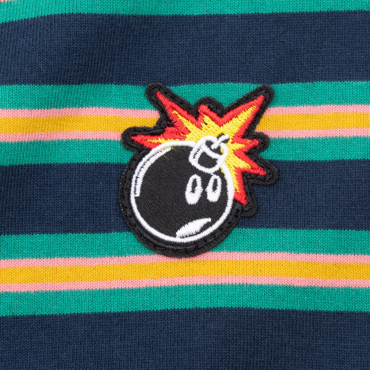 Close-up of The Hundreds bomb logo patch on navy, green, yellow, and pink striped fabric