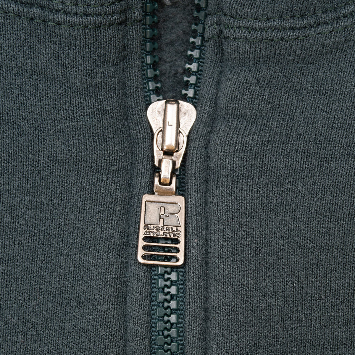 Close-up of Russell Athletic branded metal zipper pull on dark green sweatshirt
