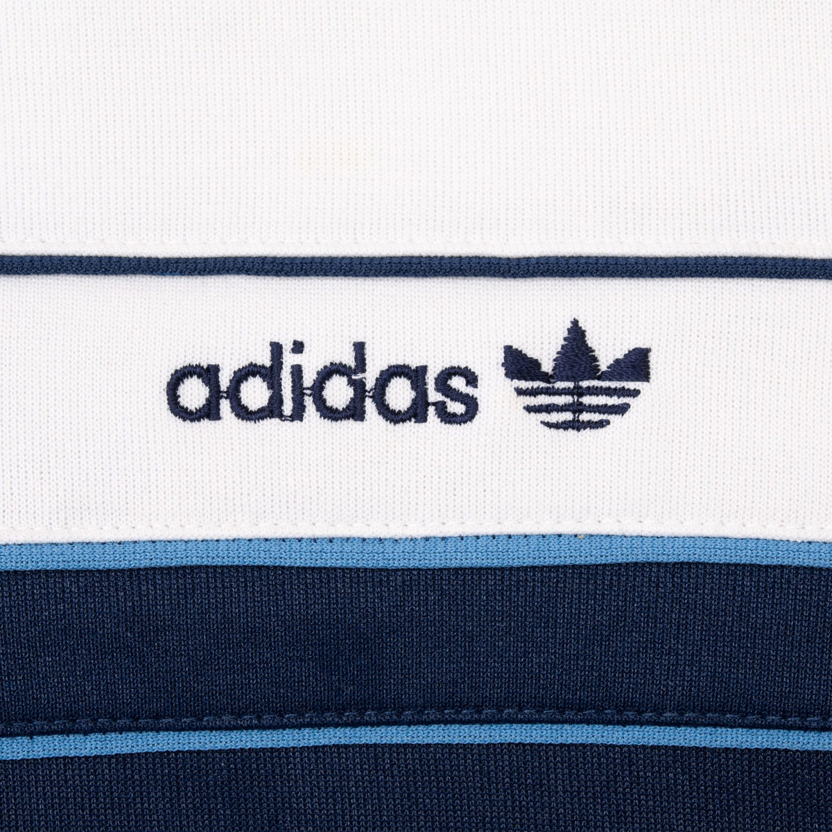 Close-up of embroidered Adidas Trefoil chest logo on white panel above blue piping