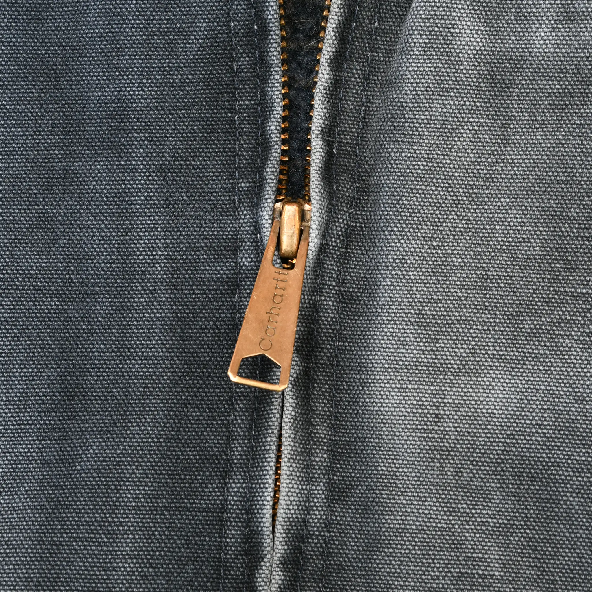 Close-up of brass-tone Carhartt zipper pull on faded gray fabric