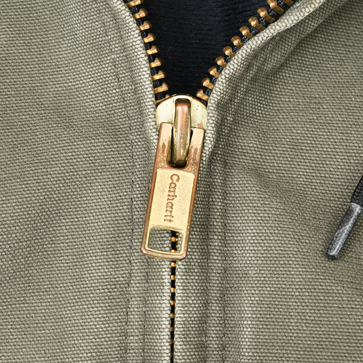 Close-up of gold-tone Carhartt zipper pull on green jacket fabric