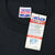 Close-up of Trench Ultra neck label and attached product hang tag on black T-shirt