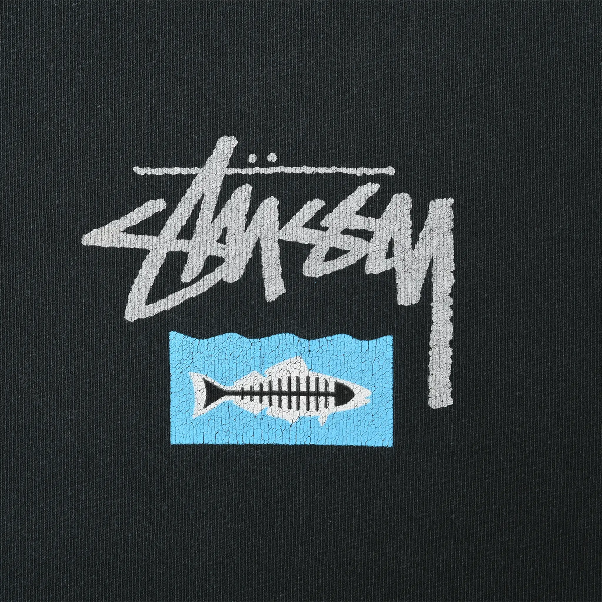 Close-up of gray Stüssy logo and fish illustration on a blue rectangle